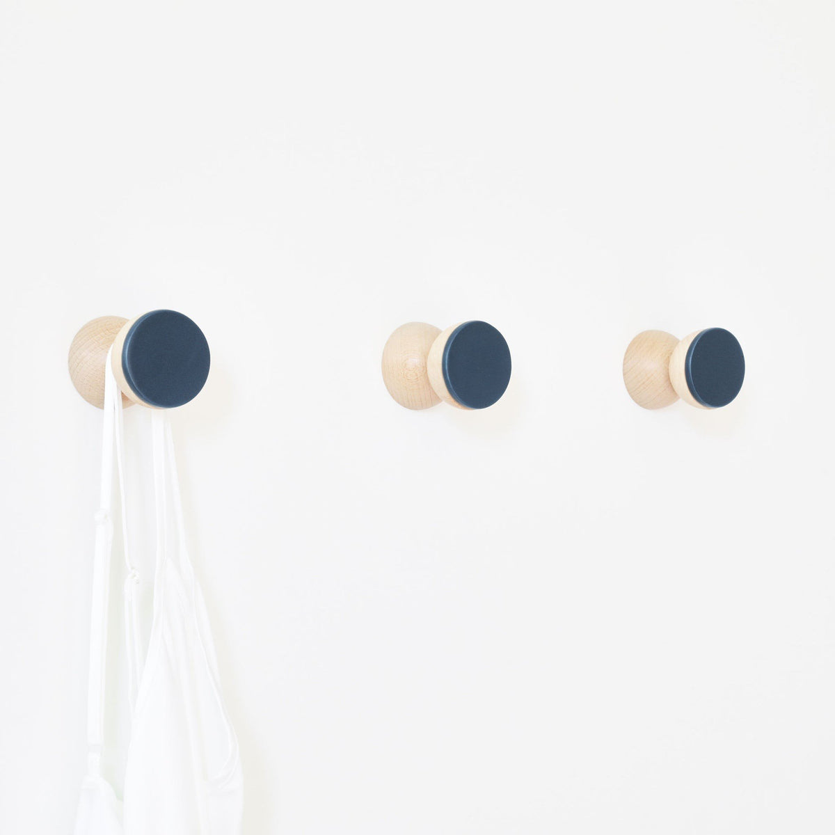 1. Trio of 5mm Paper beech wood and dark blue ceramic wall hooks holding a white bag, showcasing minimalist design on a white wall