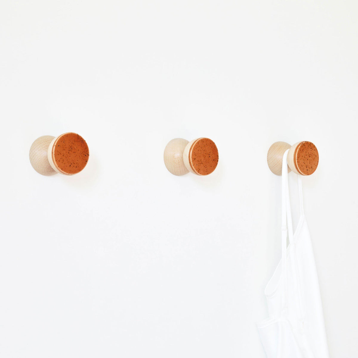 3. Studio image of three Beech Wood & Ceramic Wall Hooks in terracotta orange specks, one holding a white garment