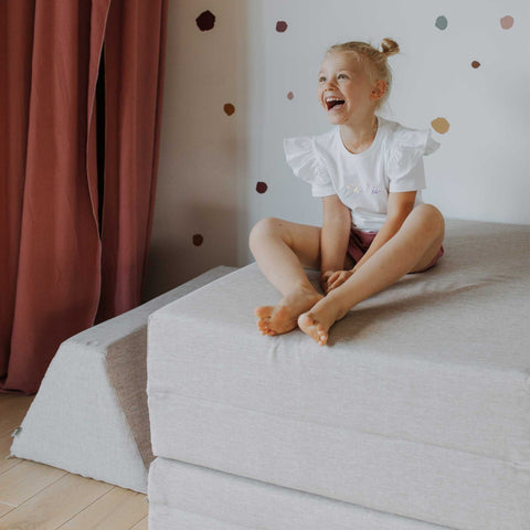 4. Smiling child sitting on stacked beige Monboxy play mattresses in a room with red curtains