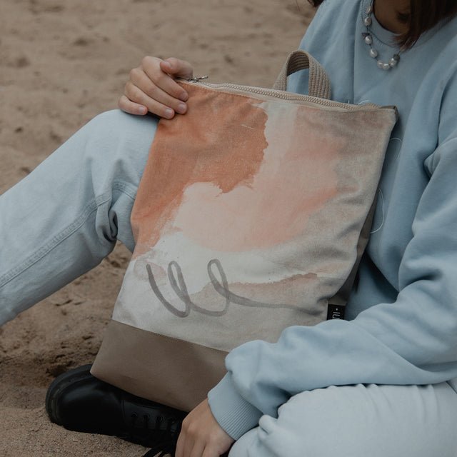1. Woman holding Muni Abstract Beige backpack with velvet and polyester design, sitting on sand