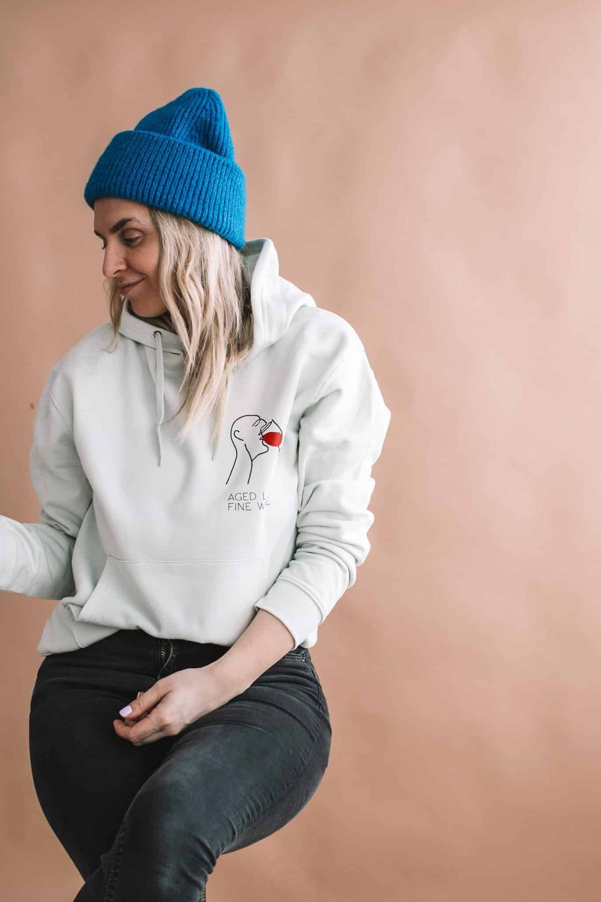 2. Woman in Black Giraffe Brand 'Aged Like Fine Wine' hoodie, light green, with blue beanie, studio setting