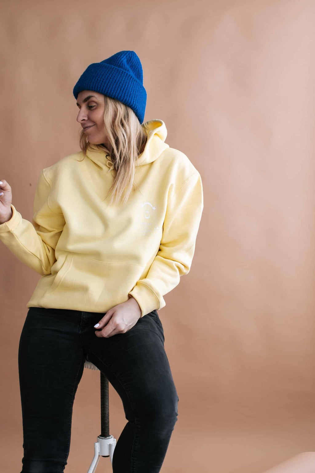 1. Woman wearing Black Giraffe Brand Note to Self Hoodie in light yellow, styled with a blue beanie, sitting in a casual pose