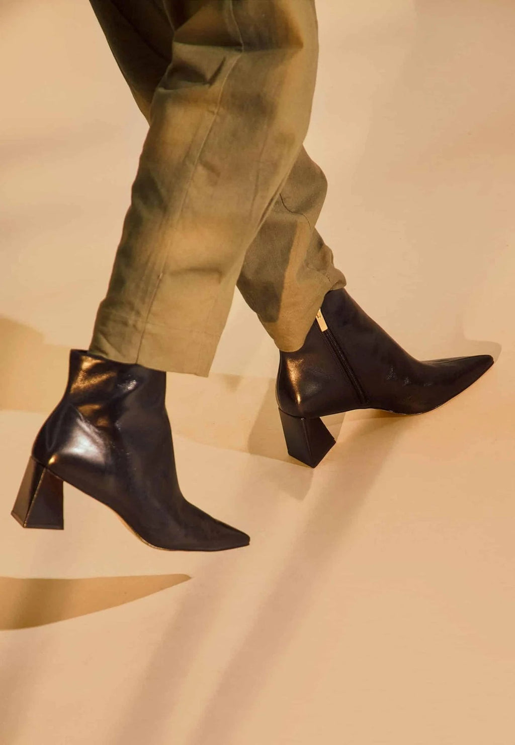 1. Woman wearing black leather ankle boots with pointed toe and block heel walking on beige background