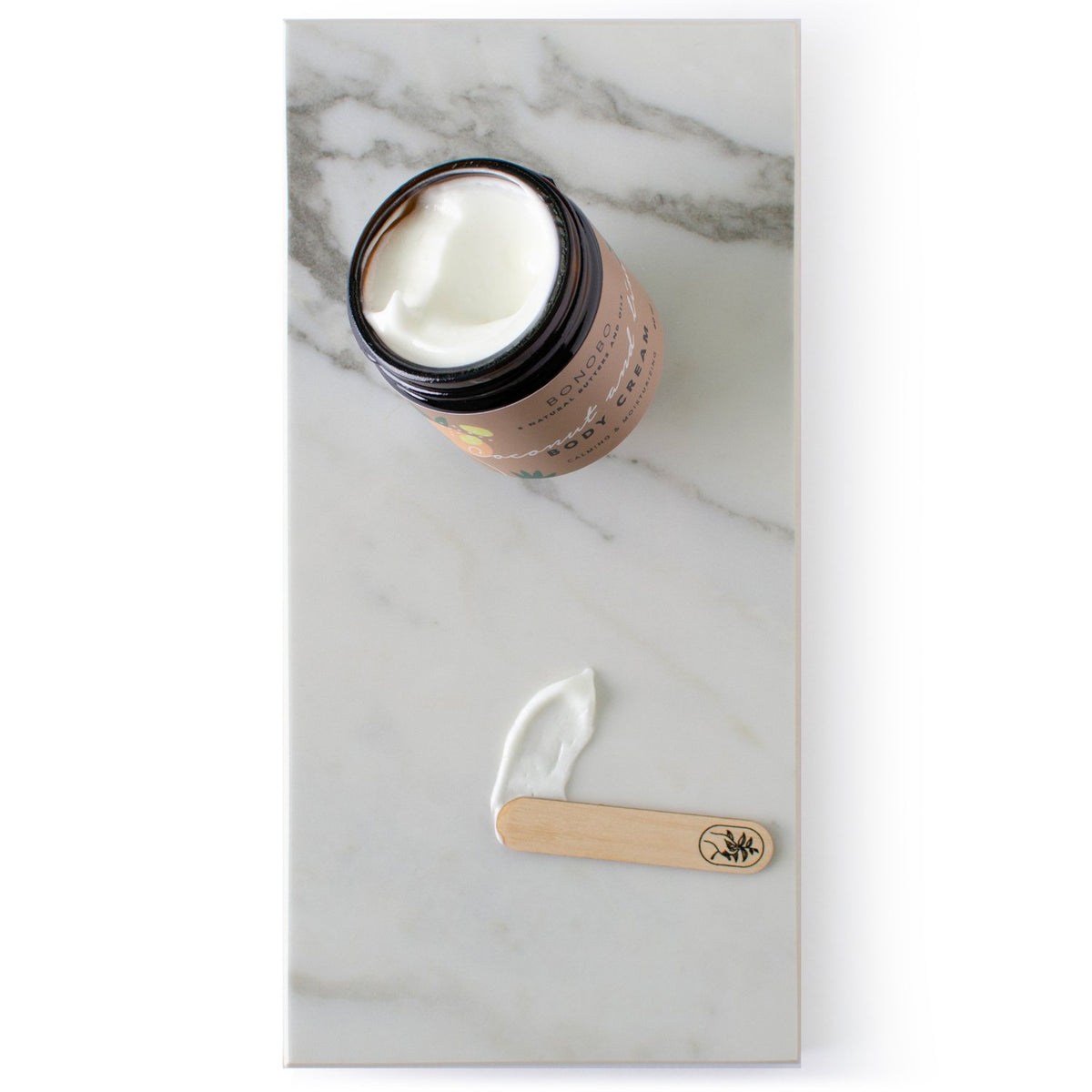 2. Flat lay of BONOBO Coconut and Lime Body Cream jar with cream on a spatula, displayed on marble