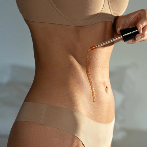 1. Woman applying shimmering body oil on abdomen with dropper, wearing beige underwear, in soft natural lighting
