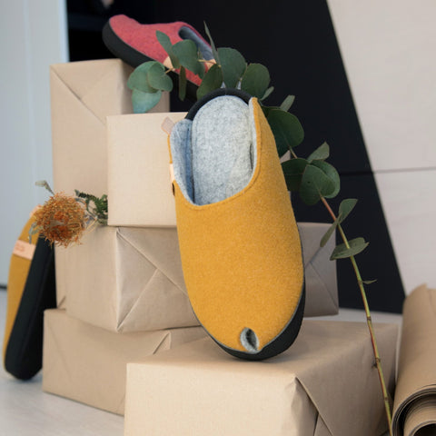 1. Lifestyle image of TOKU Brussels mustard yellow slippers displayed on a stack of boxes with greenery, highlighting cozy home style