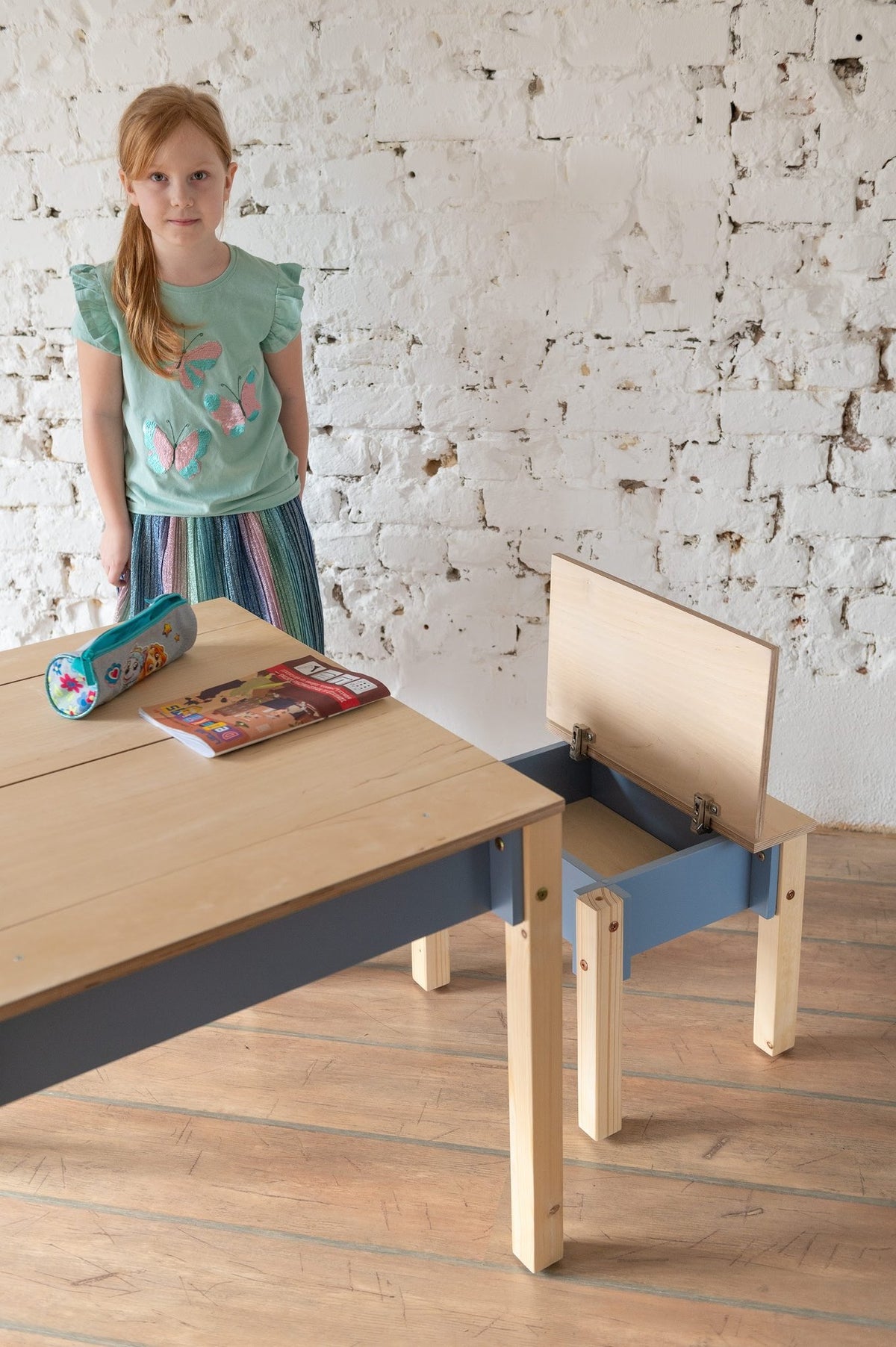 3. Girl standing next to blue and natural wood table and chair set with open storage in a room with white brick wall