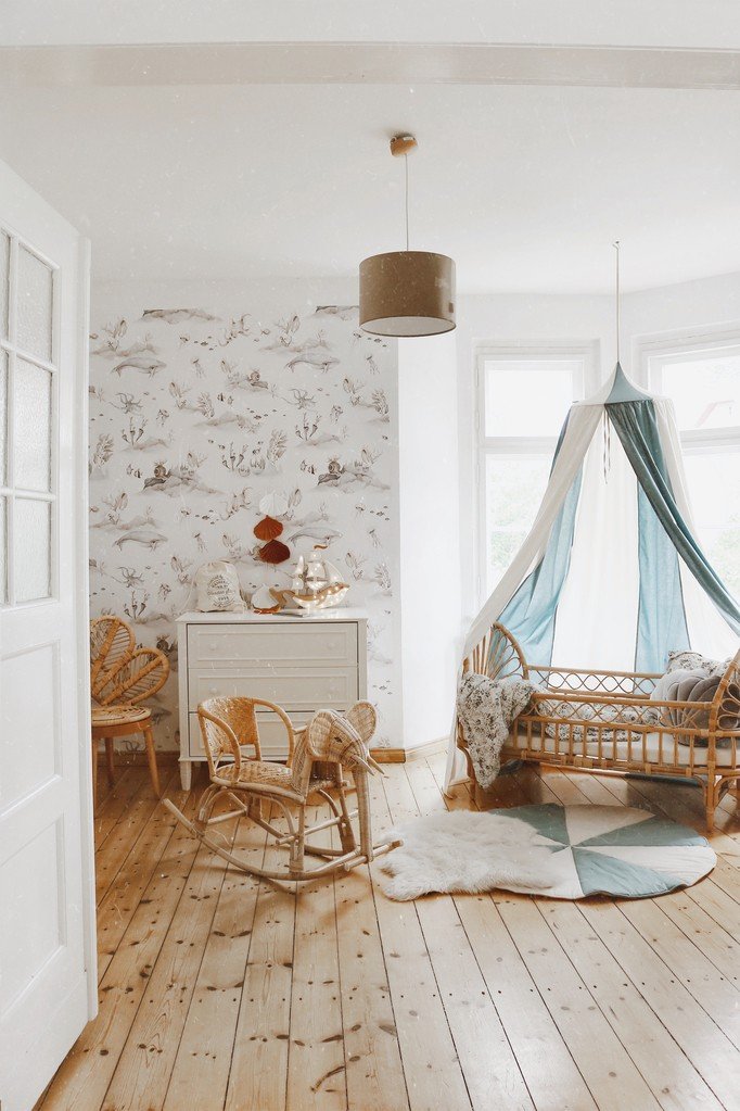 7. Nursery with blue and cream canopy over a rattan crib, wooden flooring, and whimsical wallpaper
