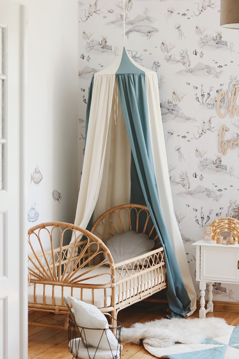 1. Blue and cream canopy hanging over a rattan crib in a nursery with whimsical wallpaper and wooden flooring