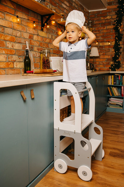 1. Child wearing chef hat on grey and white Montessori kitchen step stool in cozy kitchen