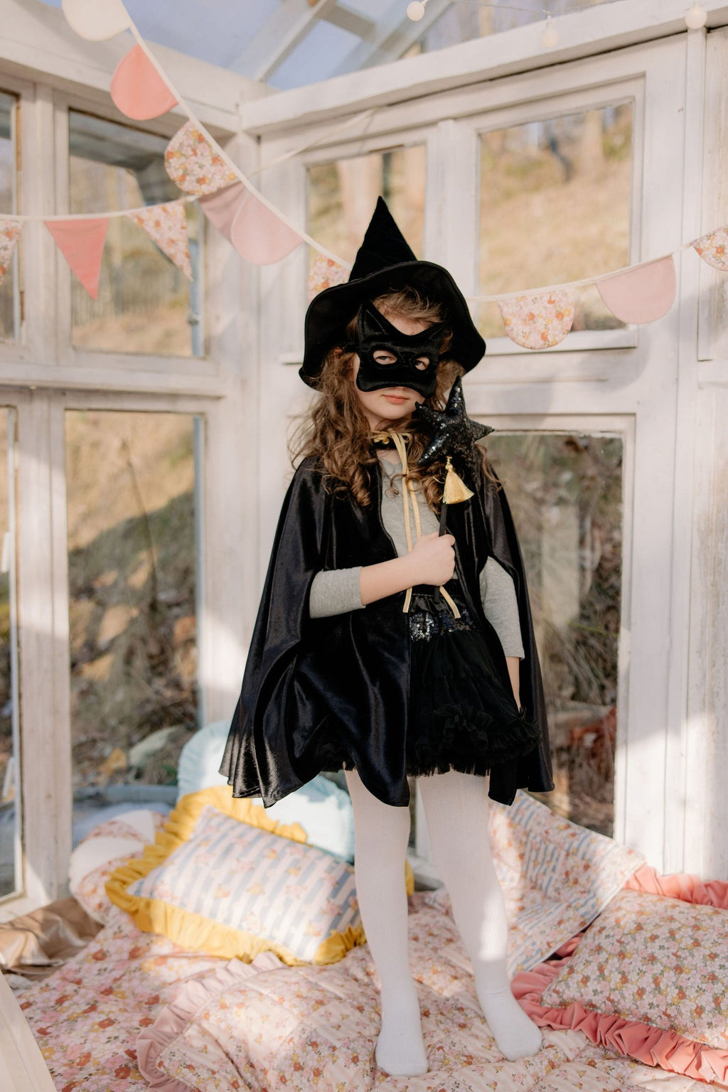 8. Child in black cat mask and cape standing in a bright room with floral decor