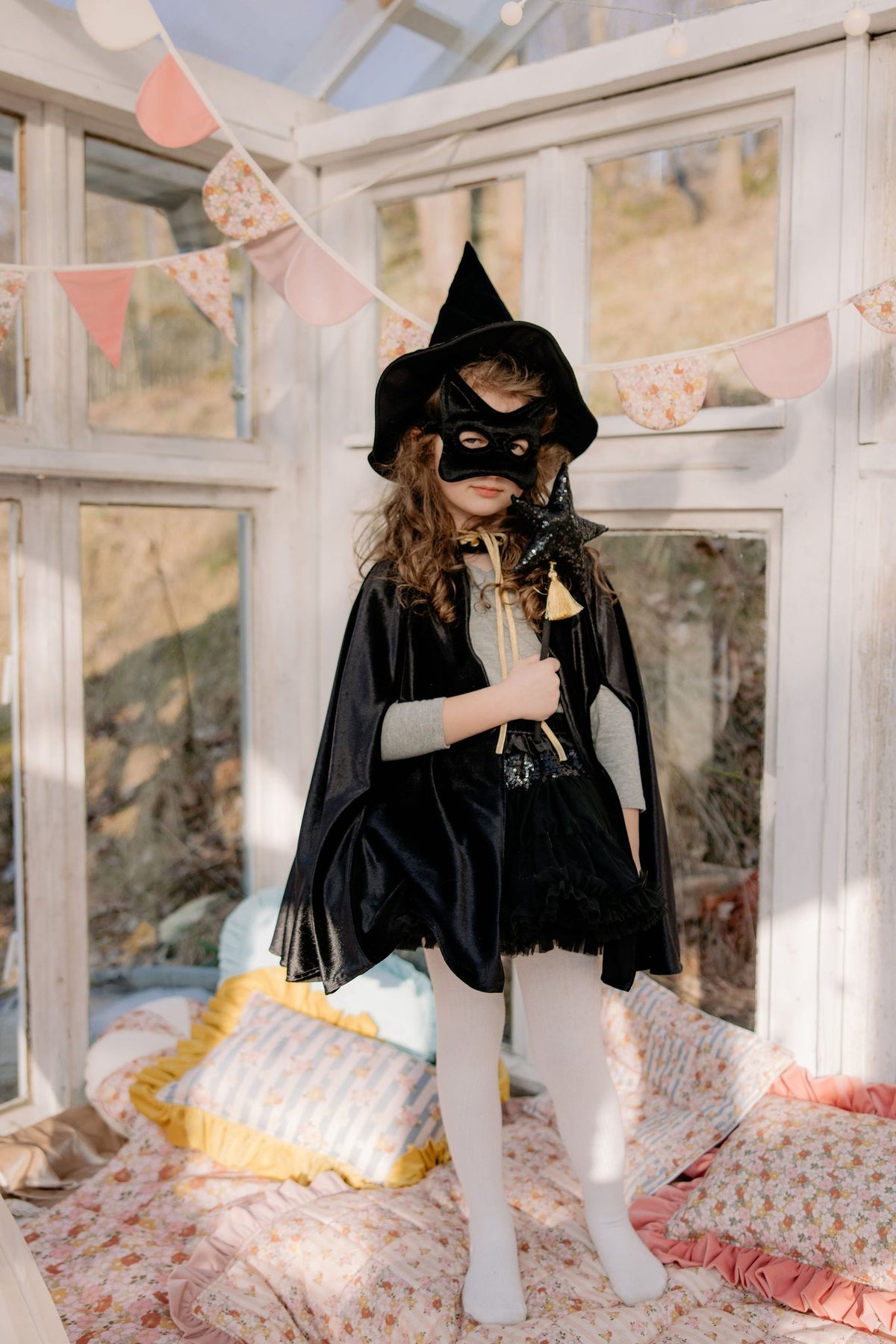 8. Child in black cat mask and cape standing in a bright room with floral decor