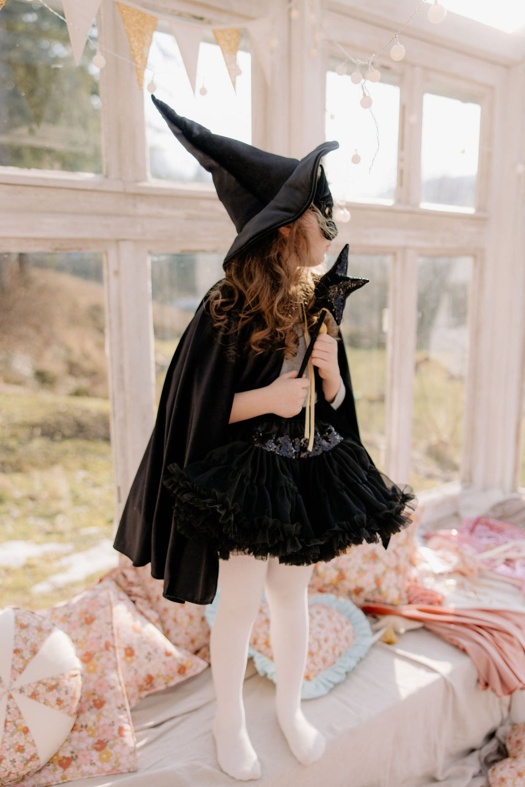 9. Child in black cat mask and cape holding a wand in a sunlit room with floral pillows