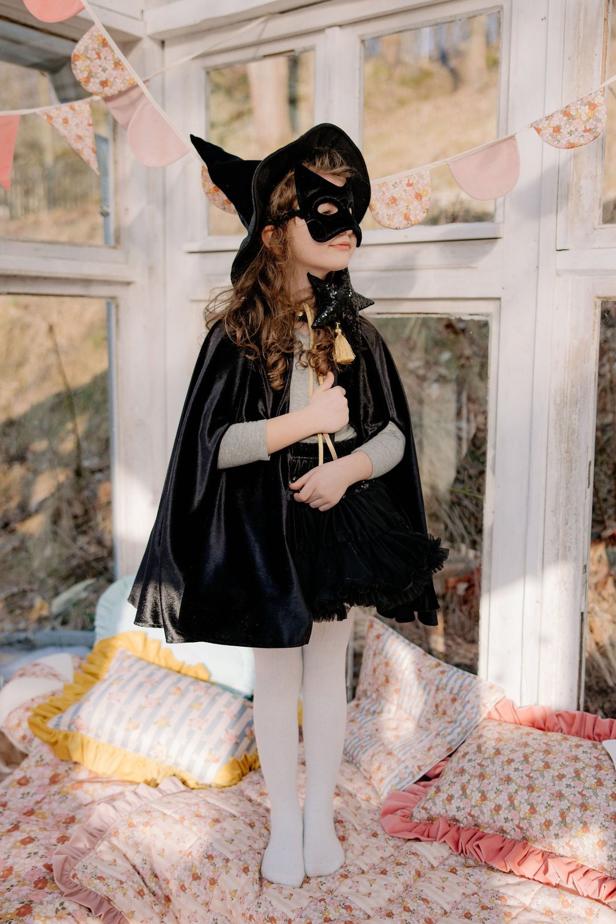1. Child wearing black cat mask and cape standing in a sunlit room with decorative pillows