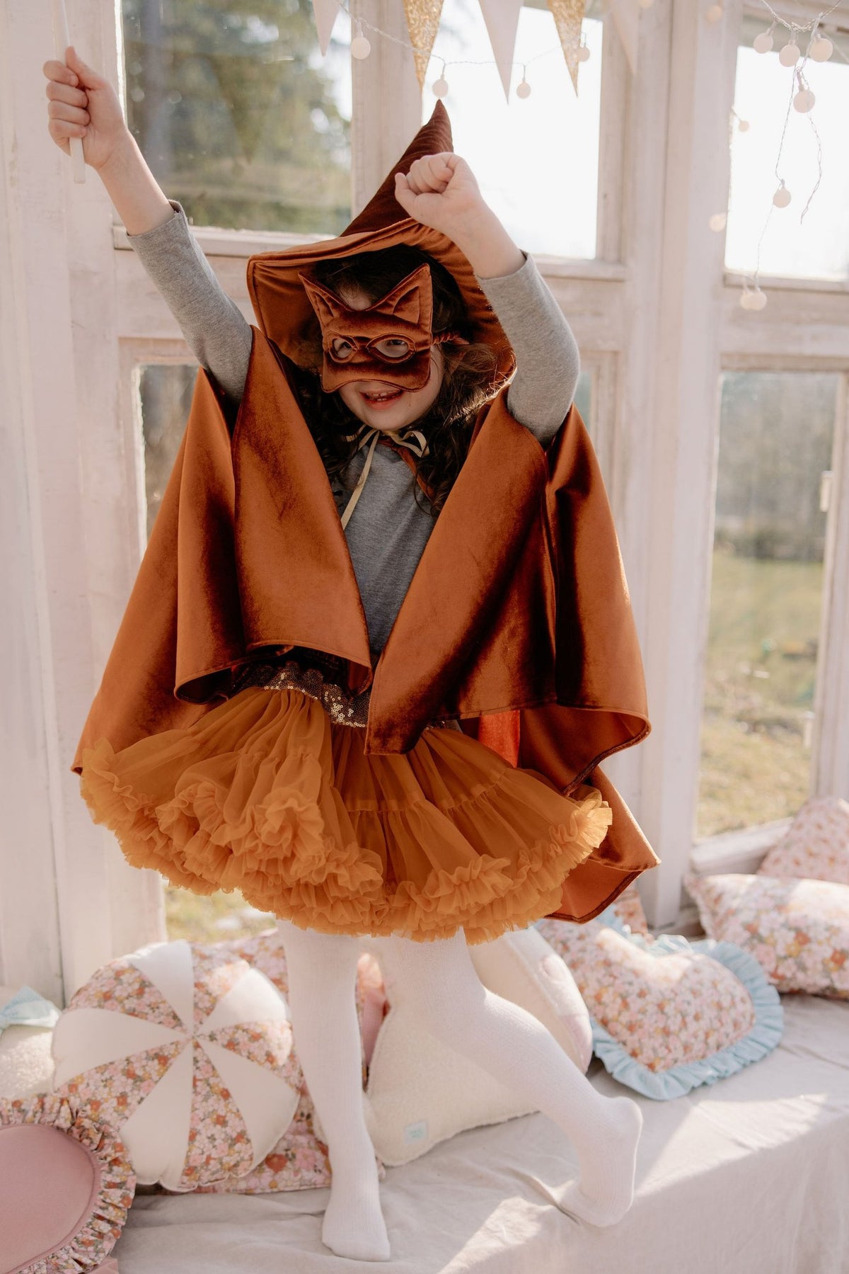 2. Girl in ginger cat mask and cape jumping on window seat with decorative pillows