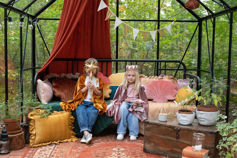 1. Two children in costume, one wearing gold sequin cat mask, sitting in a decorated greenhouse