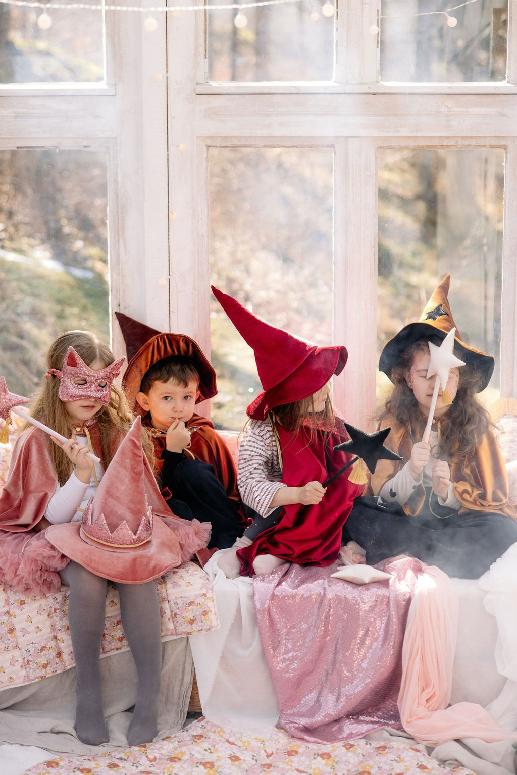 1. Group of children in costume, one wearing pink sequin cat mask, sitting on a bed in a bright room