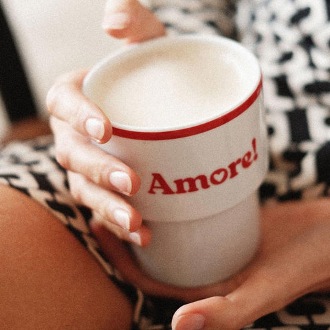 1. Woman holding Terra Mamma Amore ceramic mug with red rim and 'Amore!' text, wearing patterned clothing, in a cozy setting