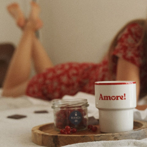 5. Terra Mamma Amore ceramic mug with red rim and 'Amore!' text on a wooden tray with berries, woman in red dress relaxing in bed