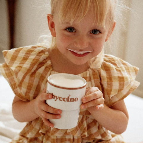 1. Child holding Terra Mamma Babyccino ceramic mug with milk mustache, wearing yellow plaid dress, smiling indoors