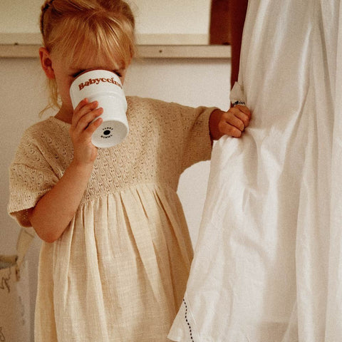 5. Child drinking from Terra Mamma Babyccino ceramic mug, wearing beige dress, standing indoors