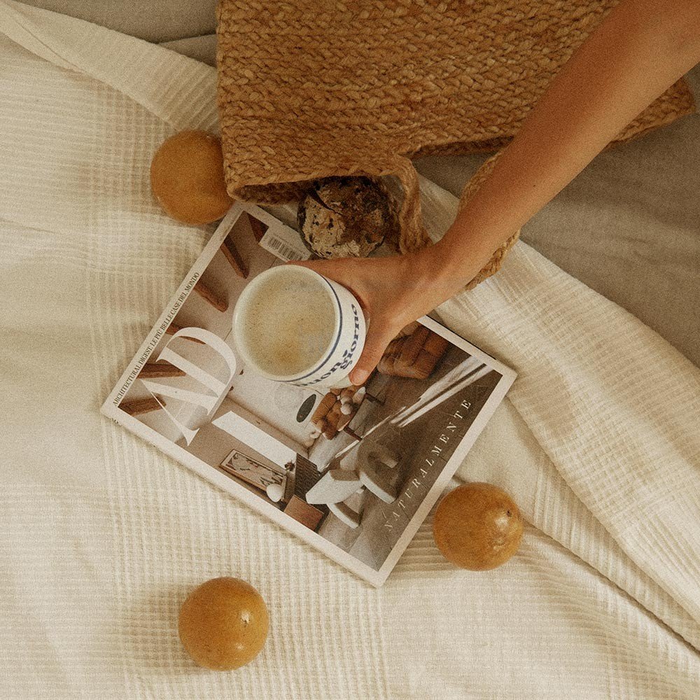 5. Hand holding Terra Mamma ceramic mug with 'buongiorno' text over magazine and fruit on bed