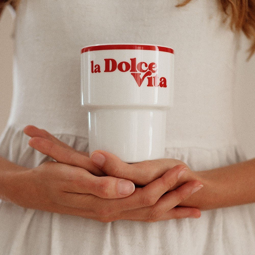 1. Woman holding Terra Mamma 'La Dolce Vita' ceramic mug with red text, wearing white dress, indoors