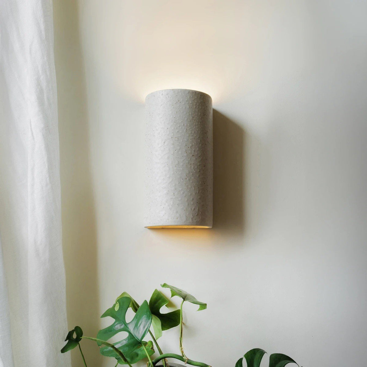 1. Beige spotted brown ceramic wall sconce on wall near window with plants, emitting soft light