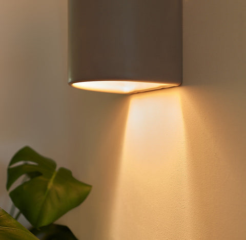 2. Close-up of taupe ceramic wall light sconce with warm light illuminating downward, next to green plant leaves