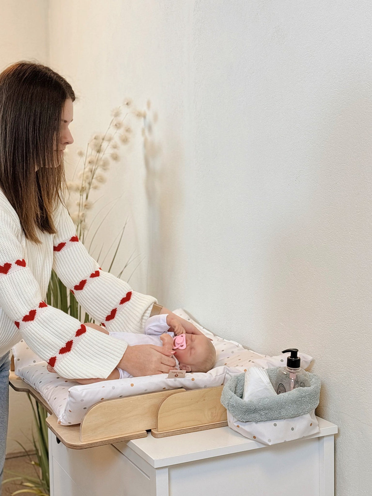 3. Woman in white sweater with red hearts changing a baby on a polka dot mat with a grey storage basket nearby