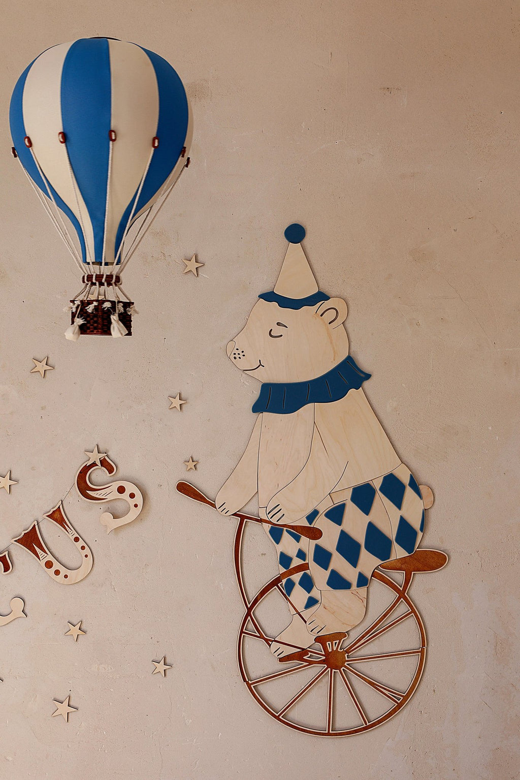 1. Wooden wall decoration featuring a teddy bear in a blue and white harlequin outfit riding a retro bike, with a blue hot air balloon and stars on a beige wall.