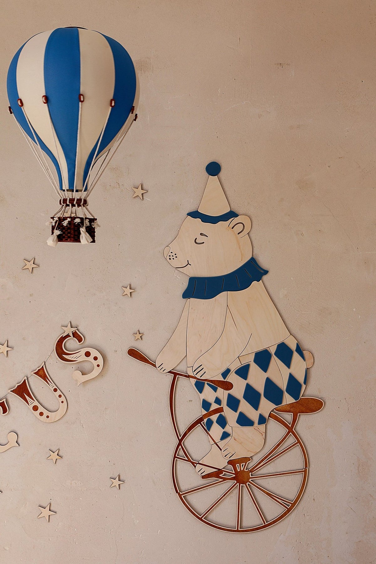1. Wooden wall decoration featuring a teddy bear in a blue and white harlequin outfit riding a retro bike, with a blue hot air balloon and stars on a beige wall.