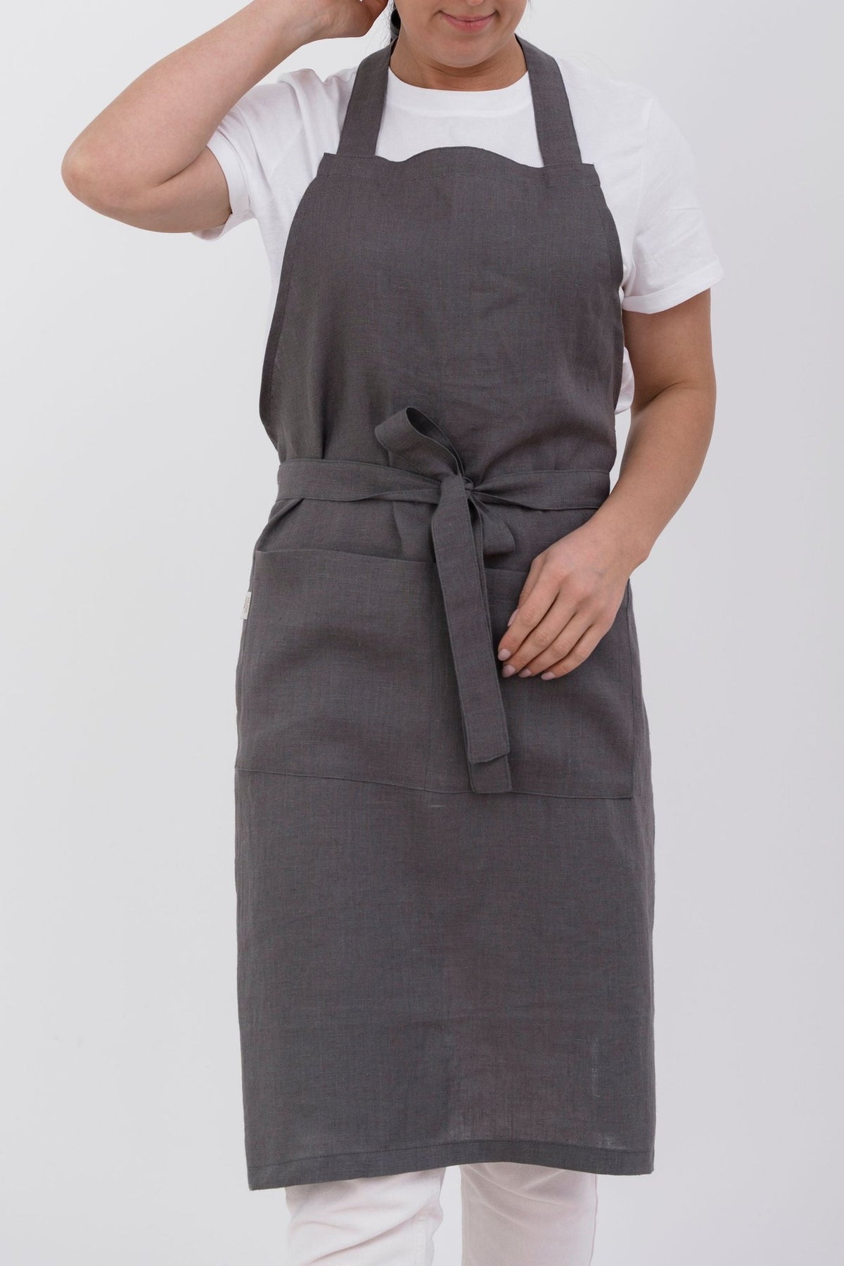 4. Woman wearing dark grey linen apron with front pocket, tied at waist, against white background