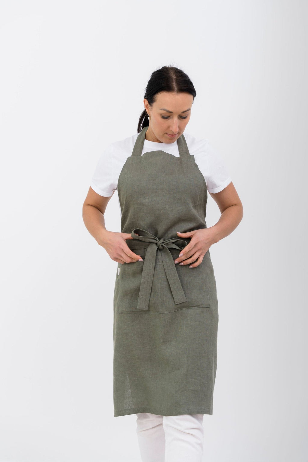 1. Woman wearing olive green linen apron with front pocket, tied at waist, against white background