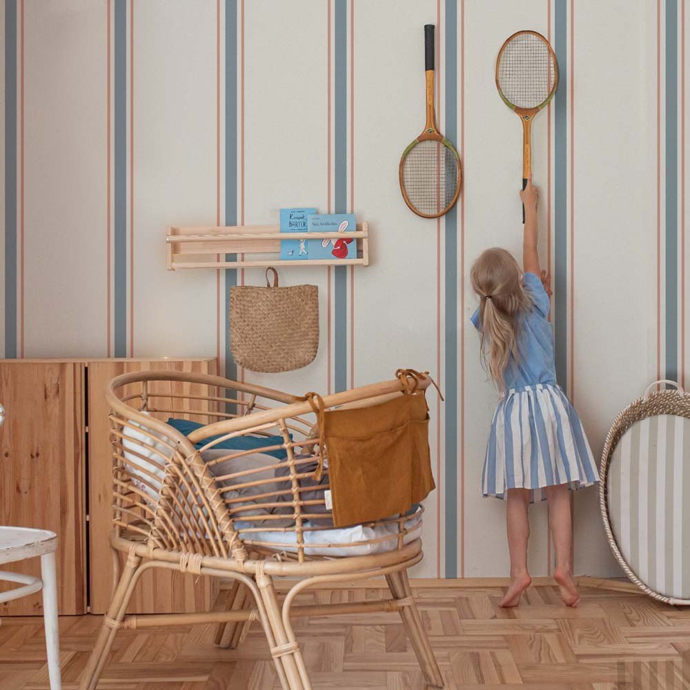 4. Girl reaching for rackets on wall with classic stripes wallpaper in linen beige, soft blue, and muted red, next to wicker furniture