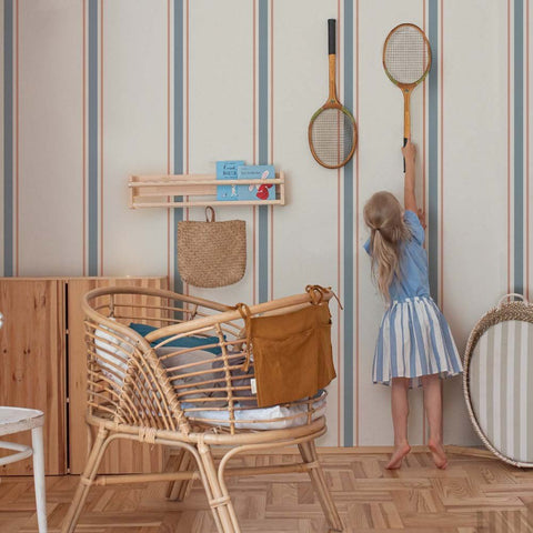 4. Girl reaching for rackets on wall with classic stripes wallpaper in linen beige, soft blue, and muted red, next to wicker furniture