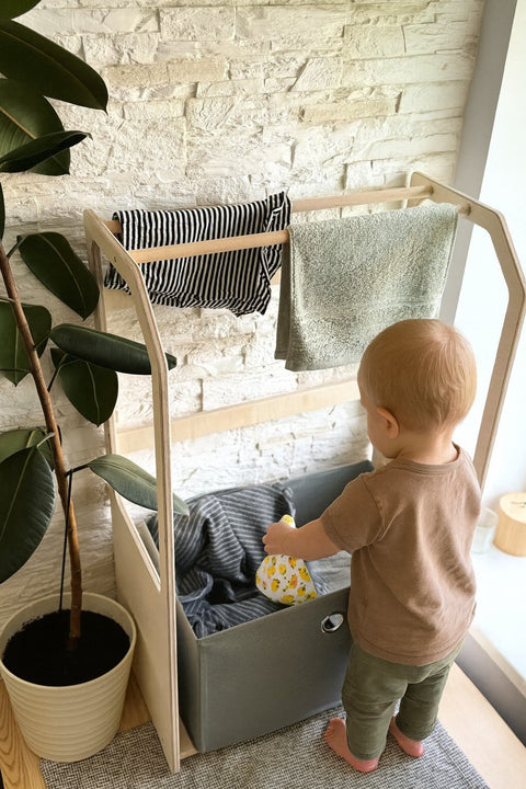 1. Toddler using Montessori wooden clothes drying rack with grey fabric basket in a bright room