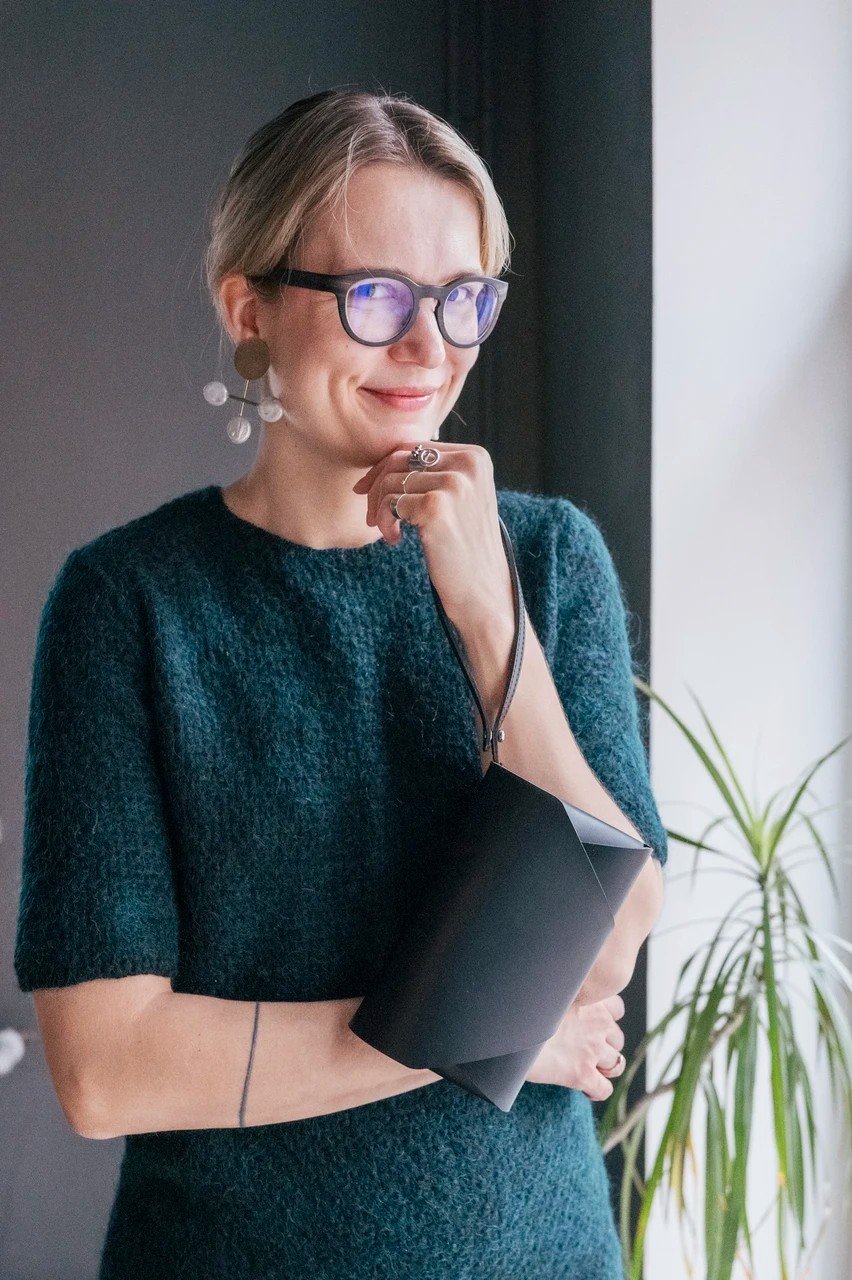 3. Woman holding black leather clutch bag with wrist strap, wearing teal sweater, smiling indoors