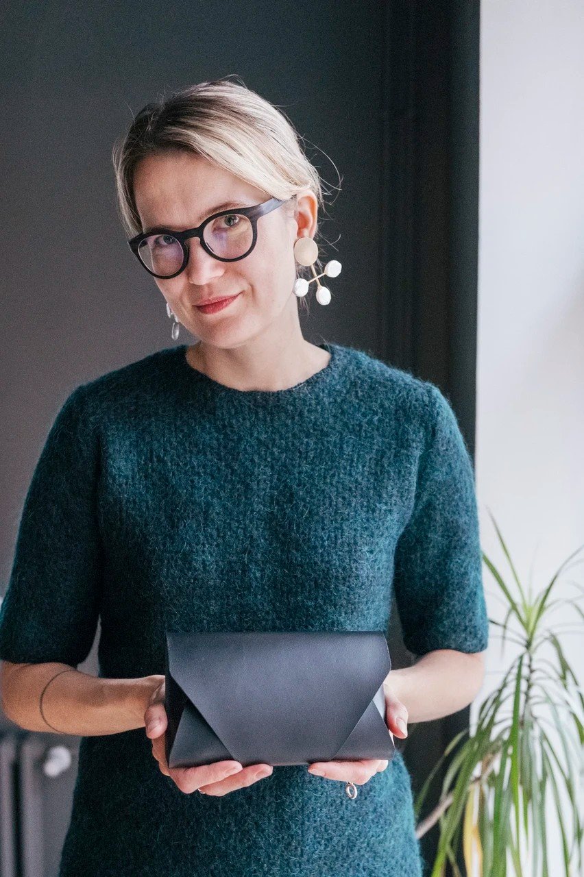 2. Woman holding black leather clutch bag in both hands, wearing teal sweater, standing indoors