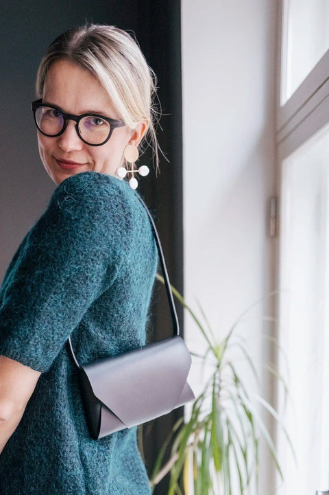 1. Woman wearing black leather clutch bag with adjustable strap over shoulder, standing by window in casual setting