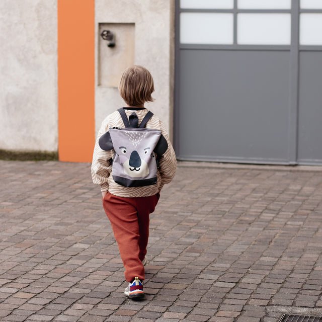 1. Child wearing Muni Koala backpack walking on cobblestone street, showcasing playful design and adjustable straps