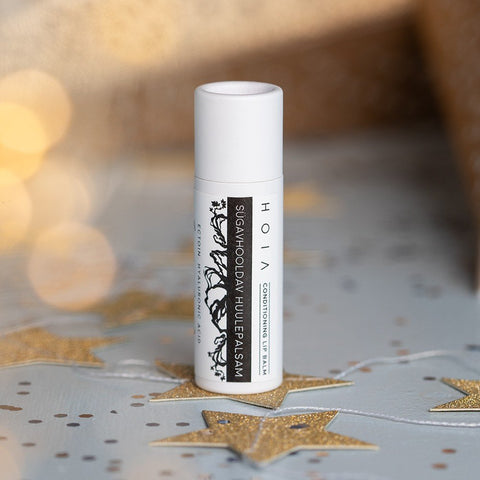 1. HOIA homespa Conditioning Lip Balm with Ectoin in white tube on festive background with gold stars, featuring black tree design and text highlighting ectoin and hyaluronic acid ingredients.