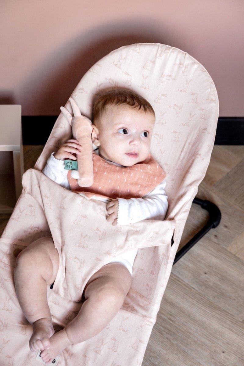 6. Baby wearing peach Trixie Baby bandana bib with dots, sitting in a pink bouncer with rabbit print