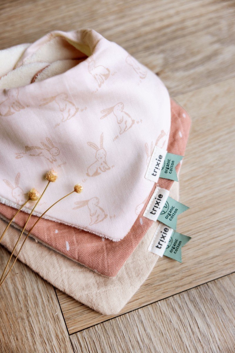 1. Three Trixie Baby bandana bibs in pink, peach, and beige with rabbit print and solid colors, laid on wooden surface