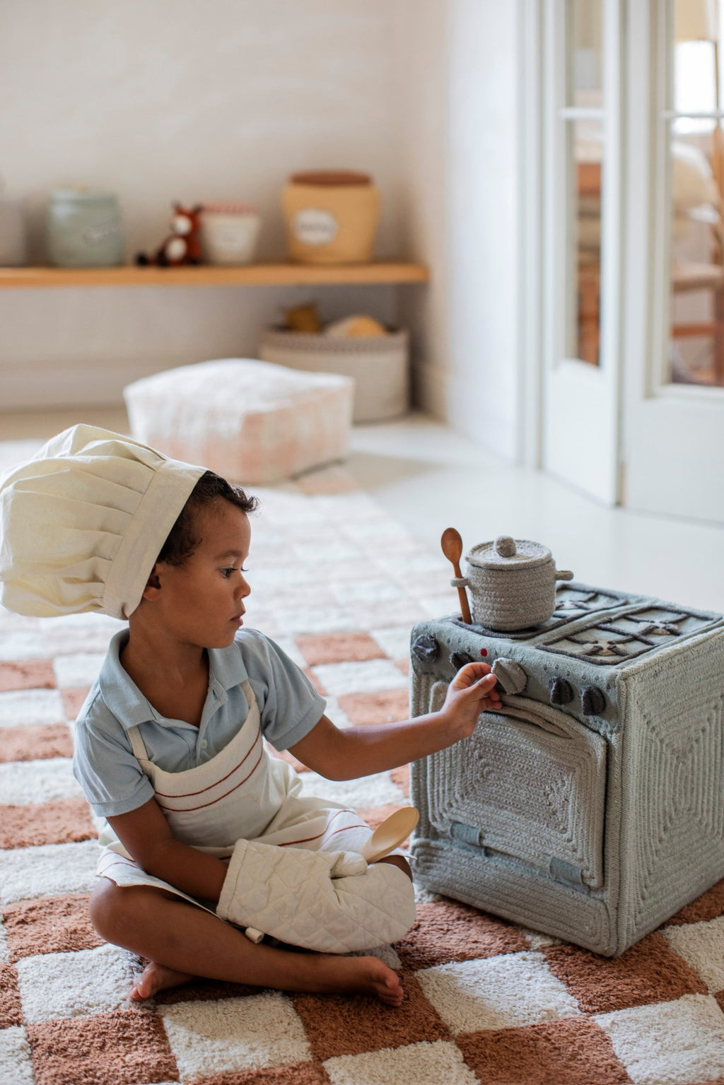 1. Child wearing chef hat and apron playing with Lorena Canals blue sage textile toy stove in cozy room