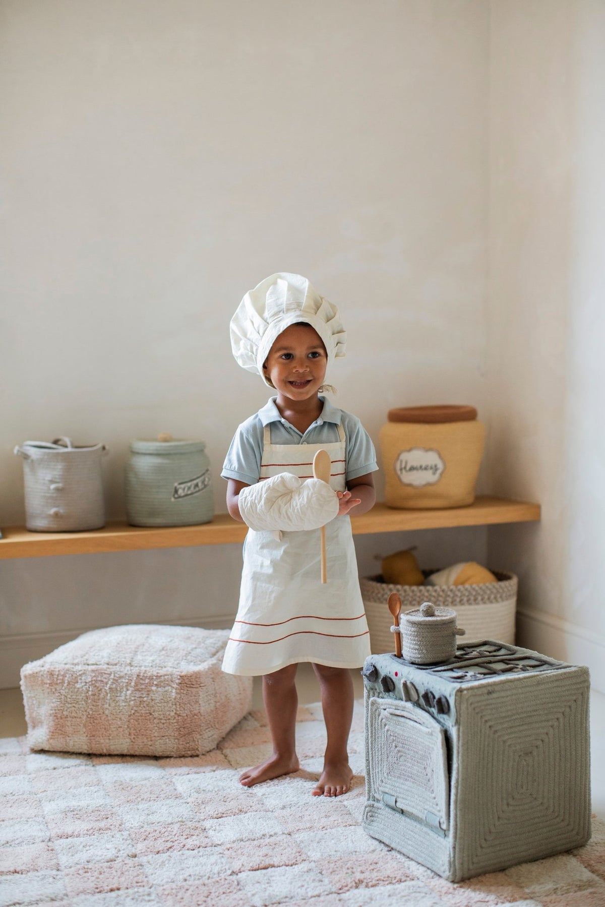 4. Smiling child in chef outfit standing next to Lorena Canals blue sage textile toy stove