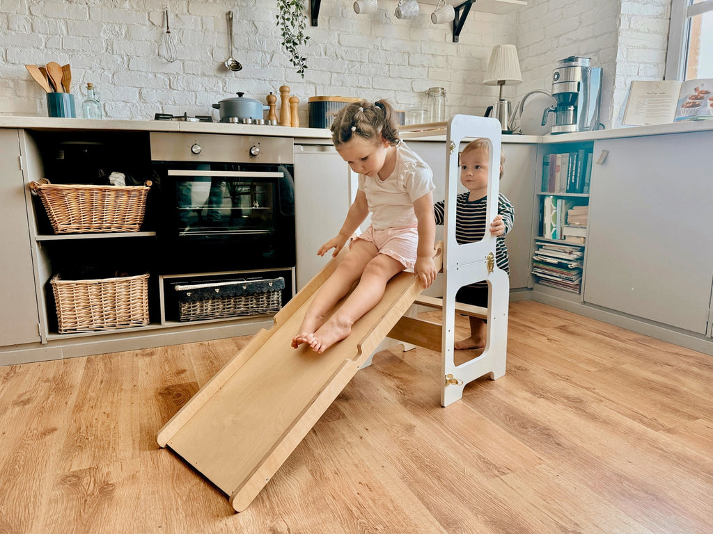 1. Two children playing on a wooden slide attached to a white learning tower in a kitchen setting