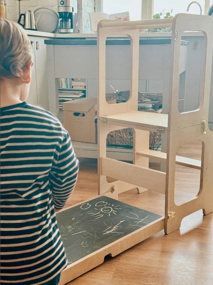4. Child standing near a wooden learning tower with a blackboard in a kitchen