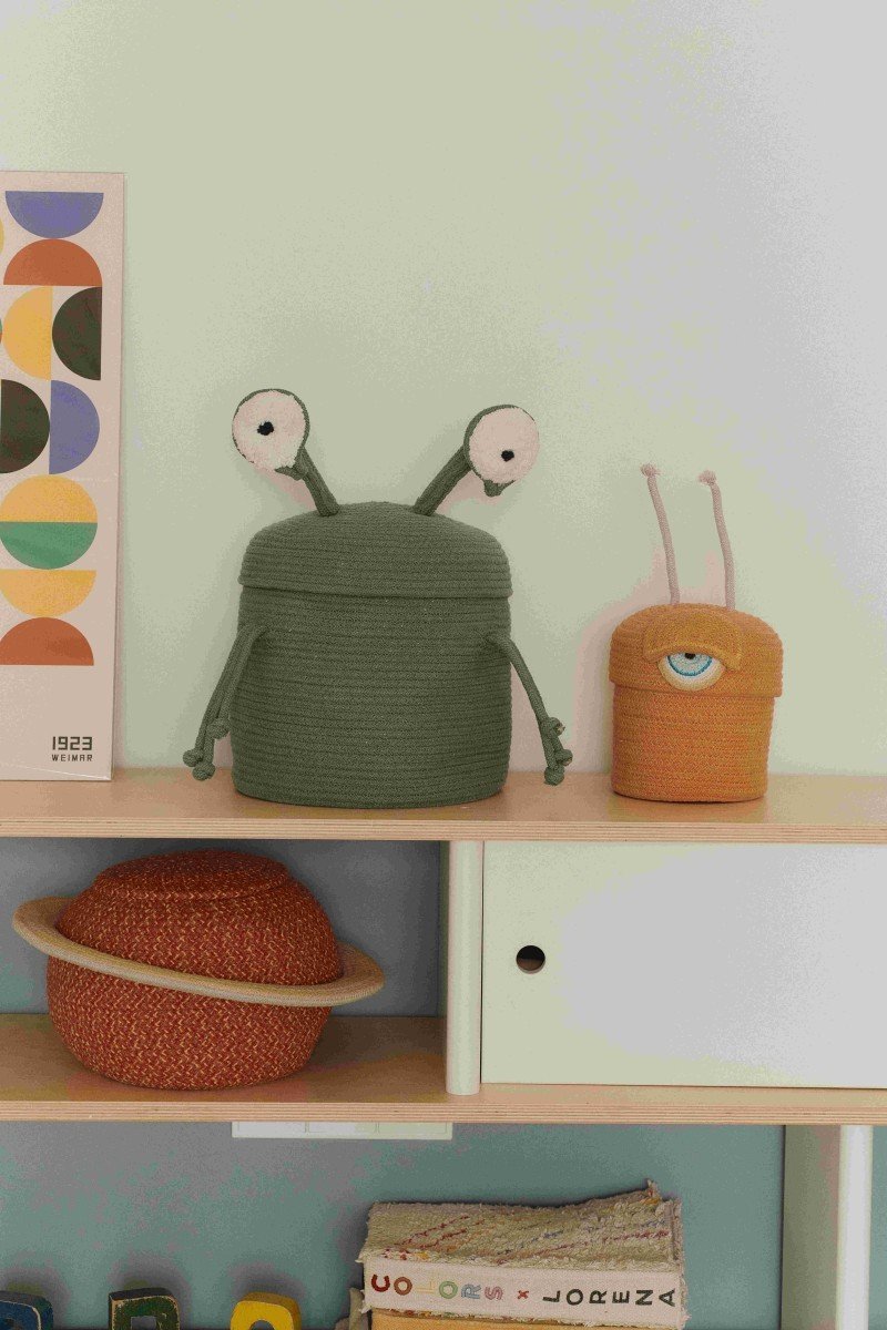 2. Green alien basket with white eyes and arm-like strings on a shelf, next to an orange basket with a single eye
