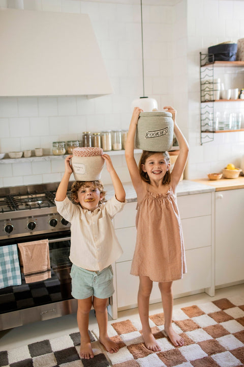 1. Two children in a kitchen holding decorative baskets, one with 'Strawberry Jam' label, showcasing playful storage solution